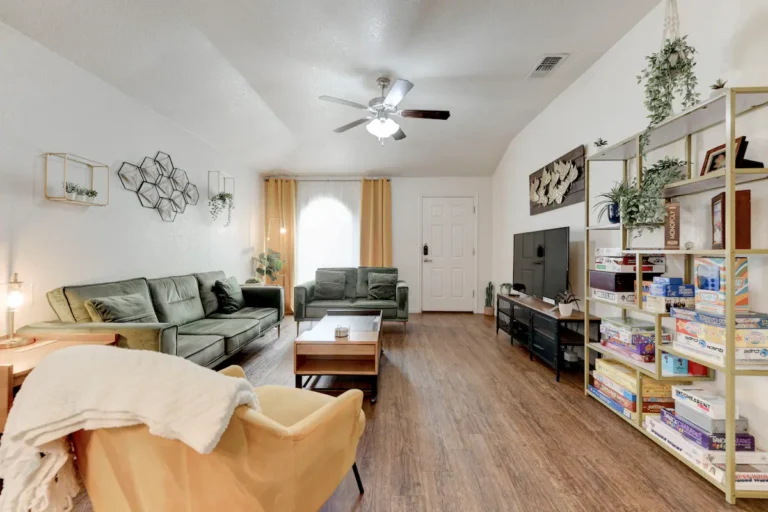 A cozy living room featuring green sofas, a yellow armchair with a blanket, a ceiling fan, and a TV on a stand. The room also includes a large bookshelf filled with board games and decor, wooden flooring, and yellow curtains near the front door.